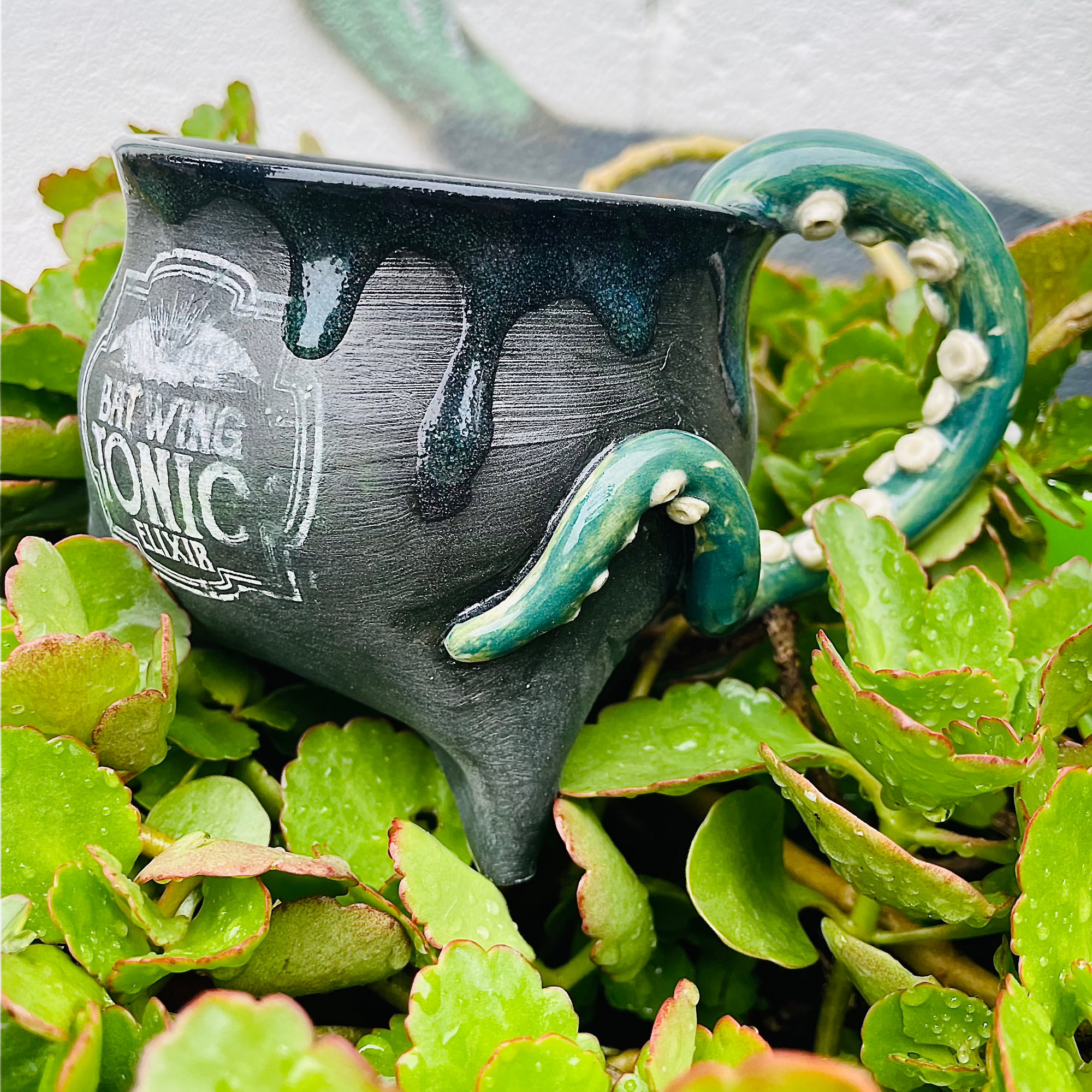 A ceramic mini cauldron with a matte black finish, featuring a handle shaped like an octopus tentacle and dripping potions, placed on a bed of green leaves.